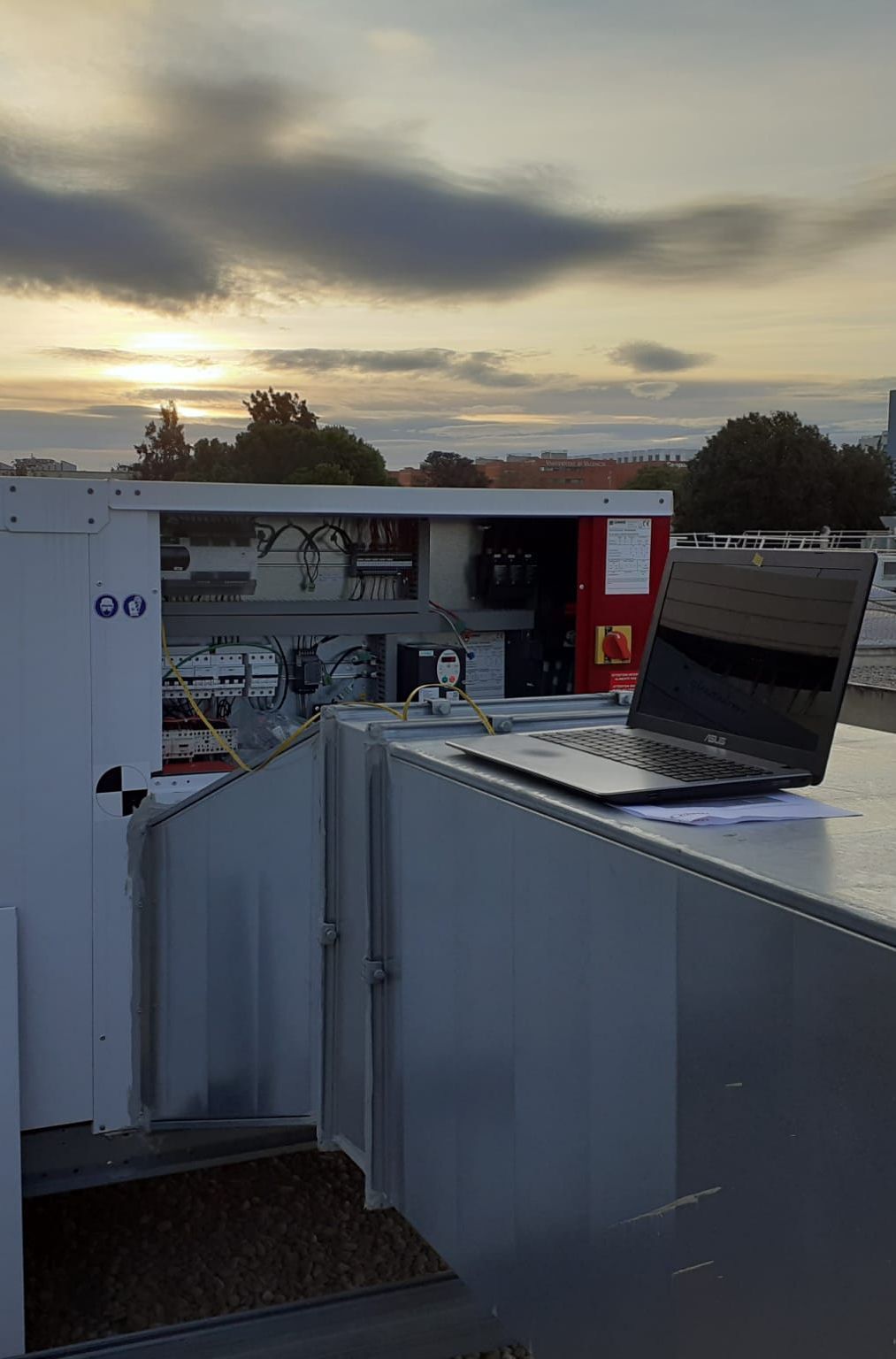Un ordenador portátil reposa sobre una superficie metálica al aire libre, al atardecer, con una unidad de control y maquinaria al fondo.