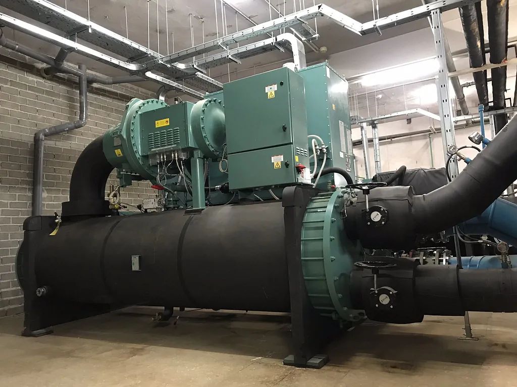 Gran unidad enfriadora industrial refrigerada por agua con tuberías aisladas de color negro en una sala de equipos mecánicos.