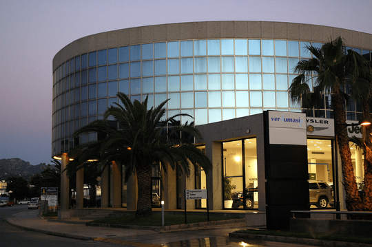 Un edificio curvo con fachada de cristal y una palmera, que funciona como concesionario de coches al anochecer.