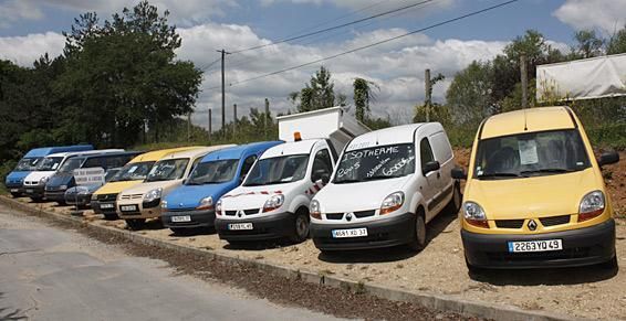 Une rangée de camionnettes de couleurs variées, garées sur un terrain gravillonné, sous un ciel nuageux.