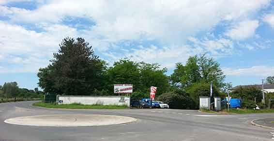 Rond-point avec un panneau, des arbres, un véhicule et un ciel bleu.