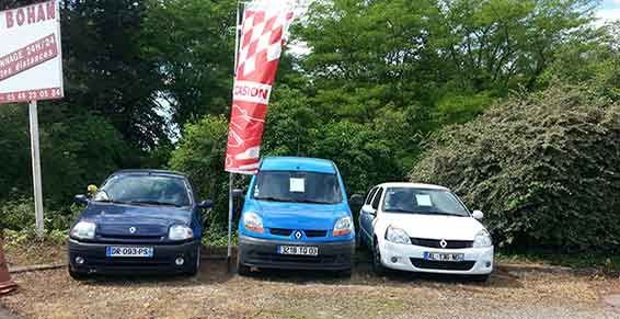 Trois voitures garées (une bleue, une camionnette bleue et une blanche) devant un espace vert. Panneau rouge et blanc visible.