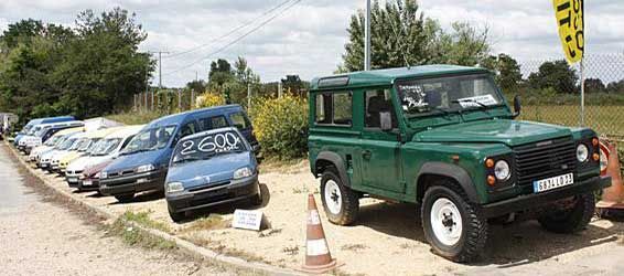 Un Land Rover vert est garé à côté d'une rangée de voitures sur un chemin de terre.
