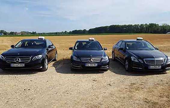 Trois Mercedes de couleur sombre sont garées sur du gravier, gyrophares allumés. Un champ et des arbres se dessinent à l'arrière-plan.