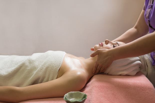 Persona recibiendo un masaje facial en un spa; manos en el cuello y la cabeza, toalla blanca, mesa rosa.