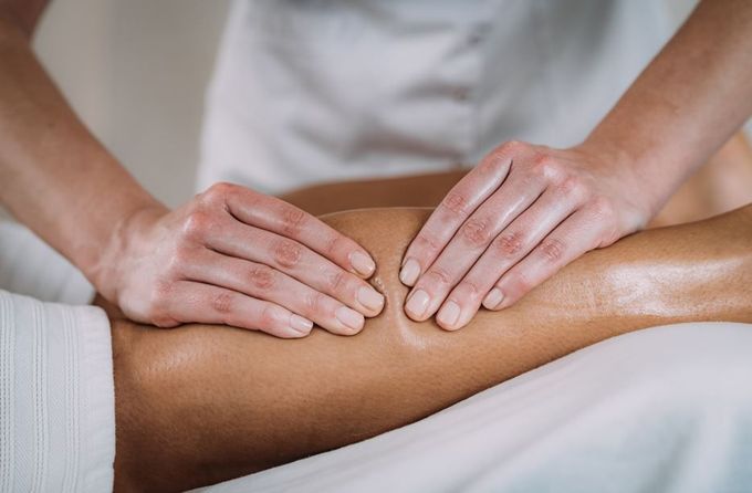 Manos masajeando la pierna de una persona. La persona yace sobre una superficie blanca, vestida con ropa blanca.