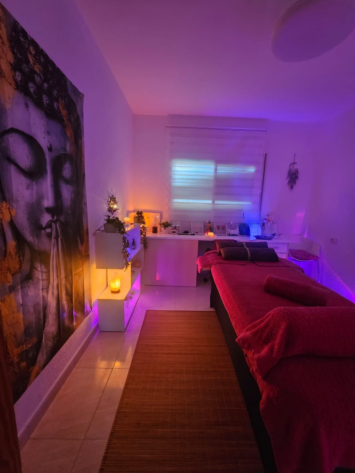 Habitación acogedora con iluminación violeta, colcha roja, cuadro de Buda en la pared y ventana con persianas.