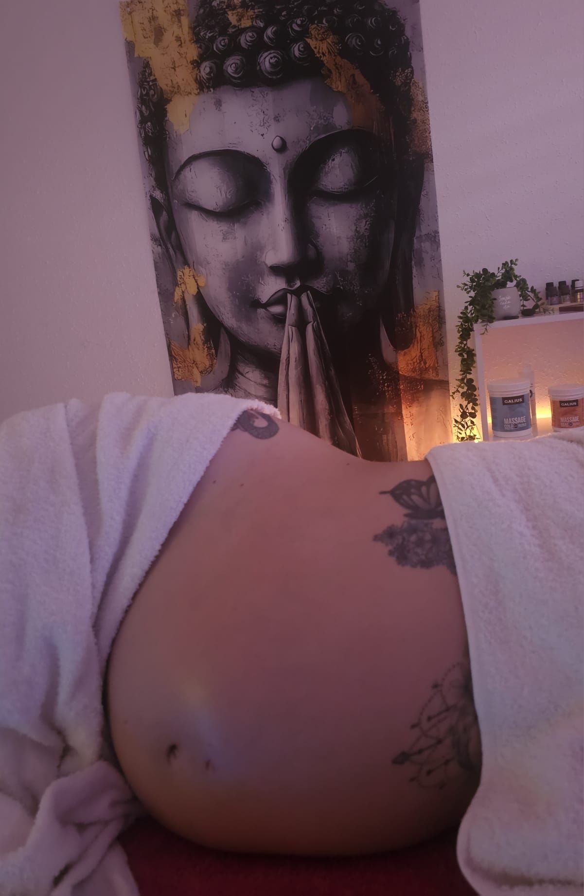 Vientre de embarazada con tatuajes en una habitación acogedora, con un cuadro de Buda en la pared y la tenue luz de las velas detrás.