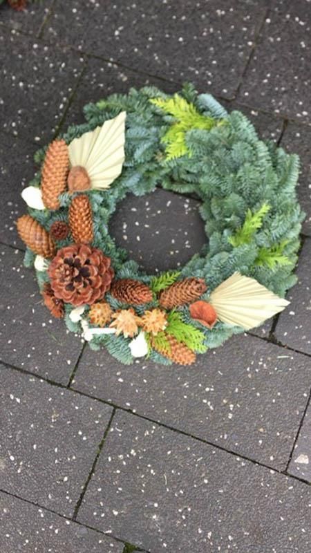 Auf einem Gehweg aus Backsteinen liegt ein Kranz mit Tannenzapfen und Blättern.