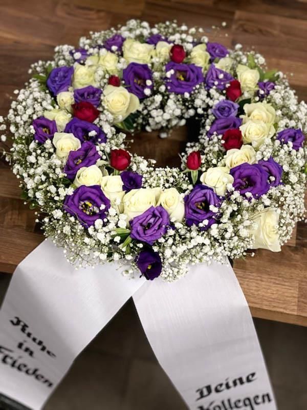 Ein Kranz aus violetten und weißen Blumen mit einer weißen Schleife auf einem Holztisch.