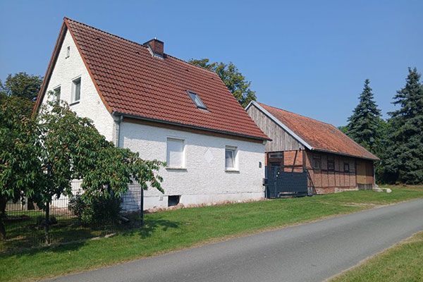 White house with red roof and attached wooden barn on a grassy roadside under a blue sky.