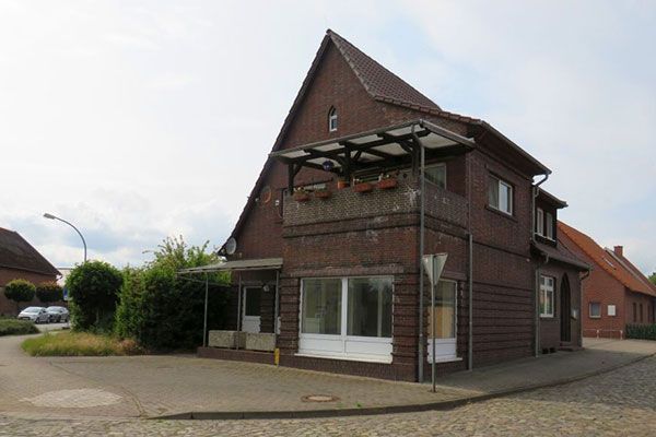 Brown brick building with a balcony. A street and green bushes are in front of the building.