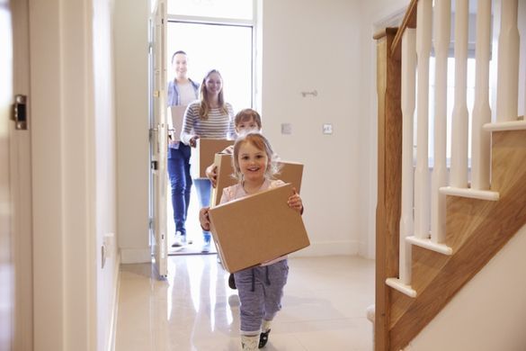 Eine Familie mit zwei Kindern zieht gerade in ein neues Haus ein. Alle kommen gerade nach- und hintereinander zur Tür rein und tragen jeweils einen Karton auf dem Arm.