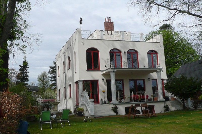 Stadtvilla mit weißer Fassade und dunkelroten Fenstern. Terrasse im Erdgeschoss mit Überdachung durch ein von zwei Säulen getragenes Dach.