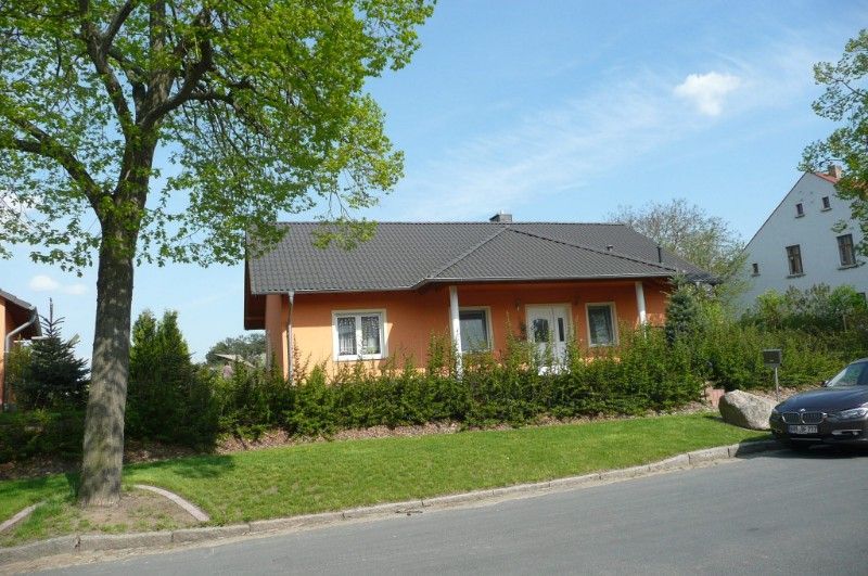 Neubau eines Einfamilienhauses mit orangefarbener Fassade, dunklem Dach und einem Vordach beim Eingang, das von zwei Säulen getragen wird.