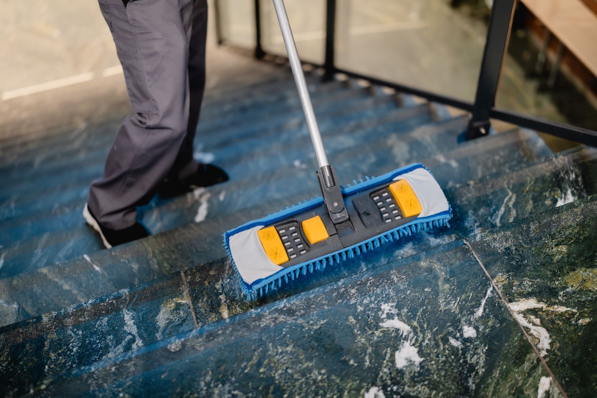 Eine Person reinigt mit einem flachen Mopp eine dunkle, gemusterte Marmortreppe.