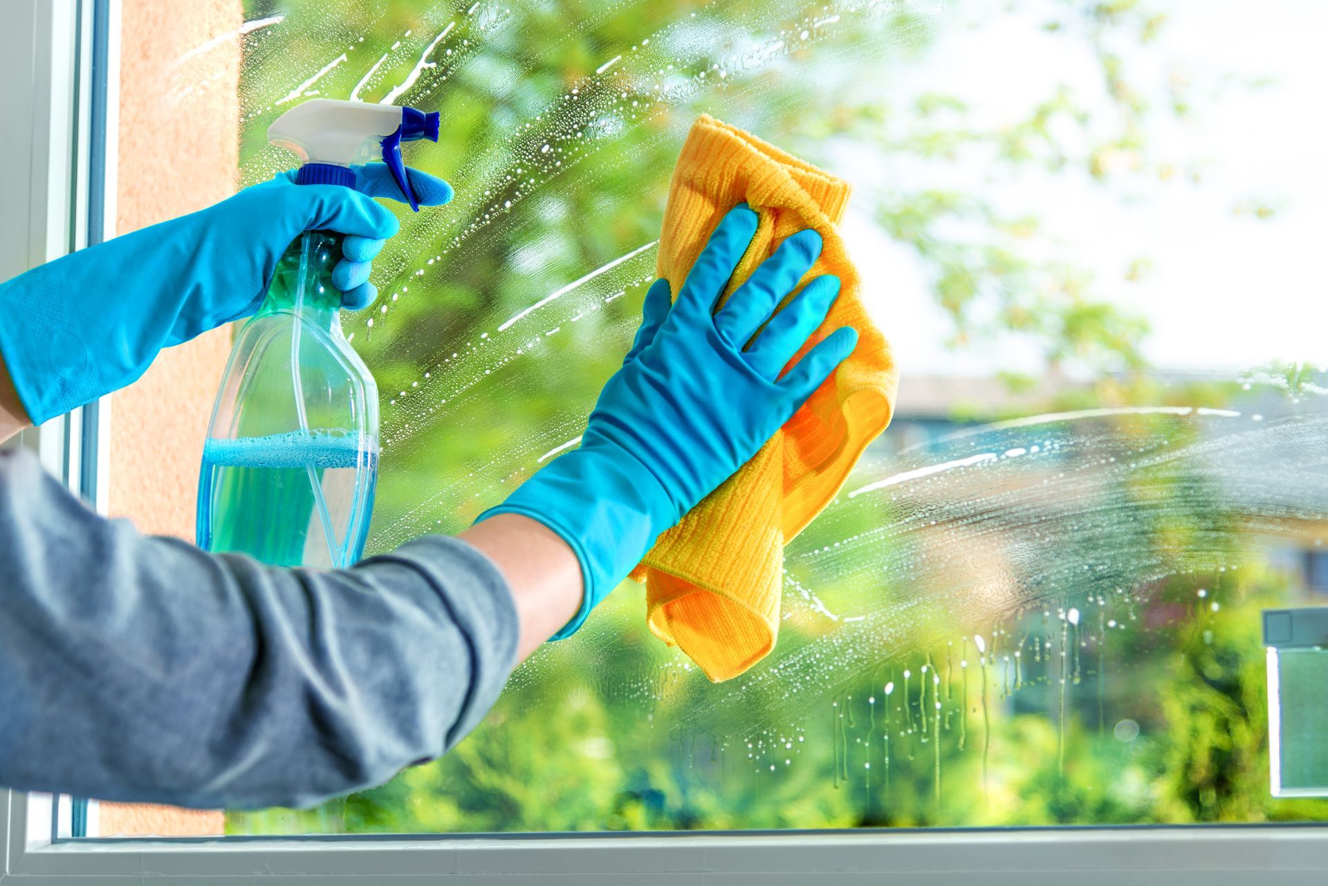 Eine Person in blauen Gummihandschuhen reinigt ein Fenster mit einem gelben Tuch und einer Sprühflasche mit Reinigungsmittel.