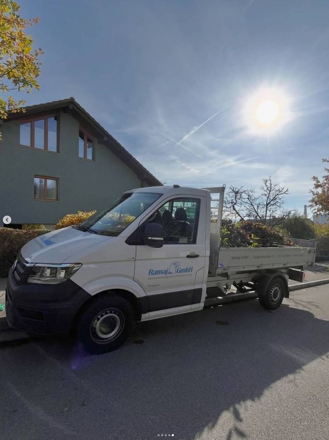 Eine Person mit Schürze und gelben Handschuhen saugt einen hellen Teppich in einem Wohnzimmer mit grauem Sofa und Hocker.