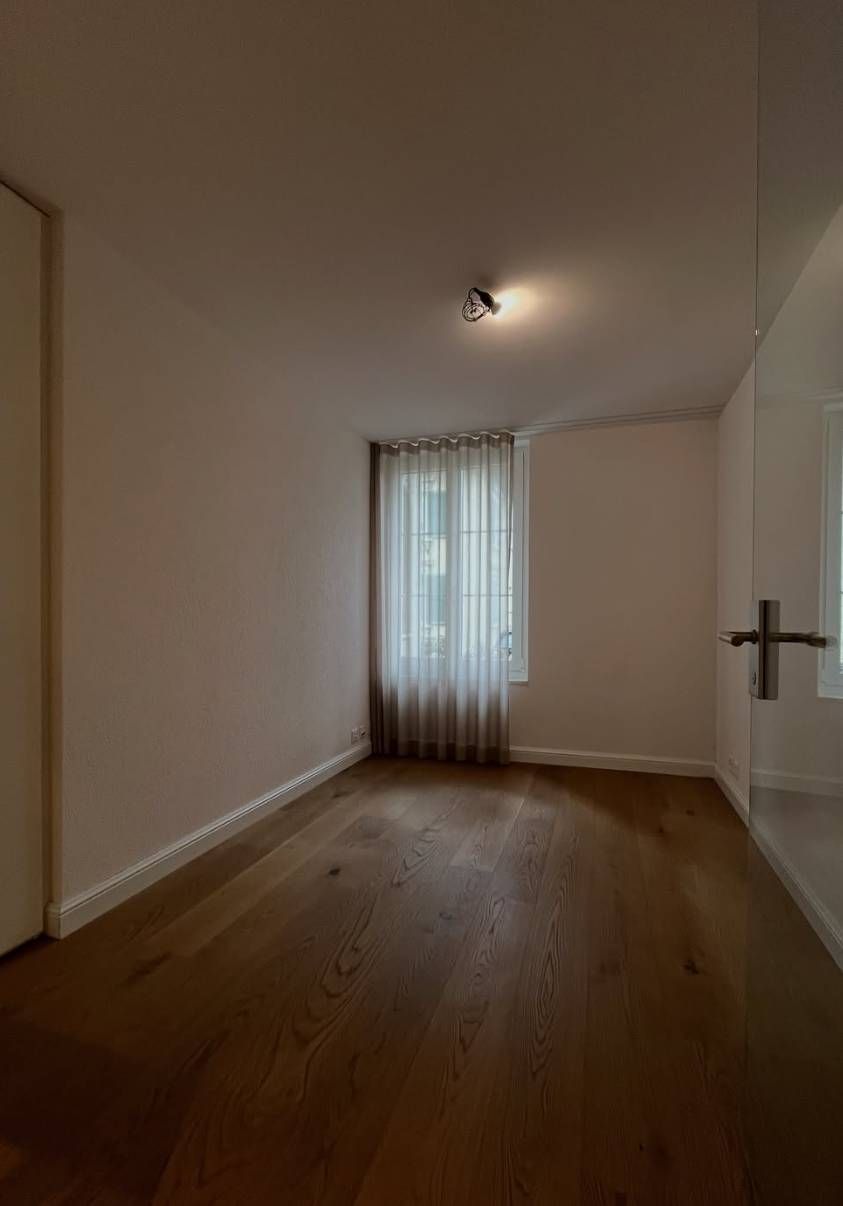 Ein leerer Raum mit hellen Holzböden, neutralen Wänden und einem Fenster, das von einem durchsichtigen weissen Vorhang verdeckt wird.