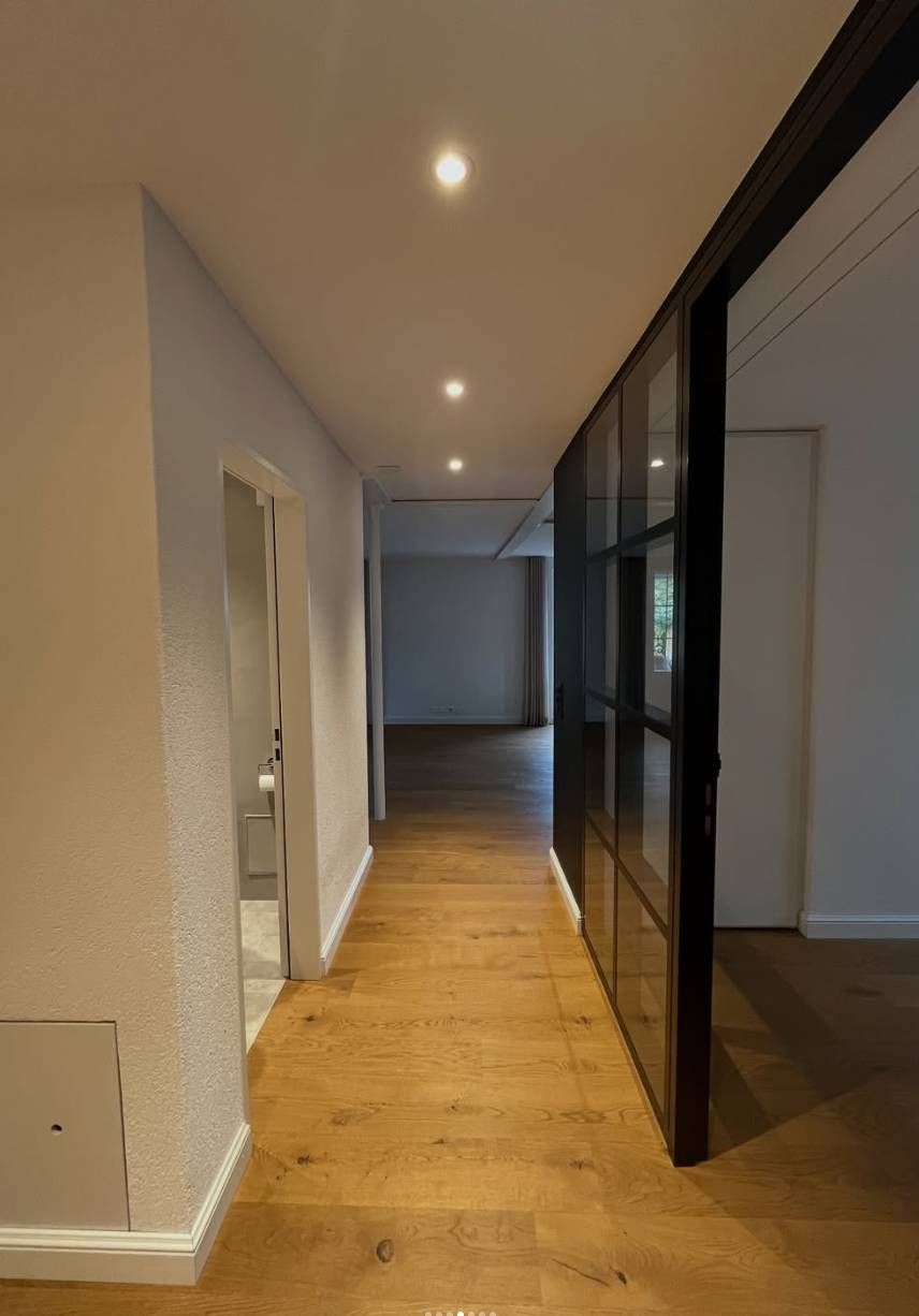 Ein Flur mit hellem Holzboden, eingelassenen Deckenleuchten, einer strukturierten Wand auf der linken Seite und einer schwarz gerahmten Glastür.