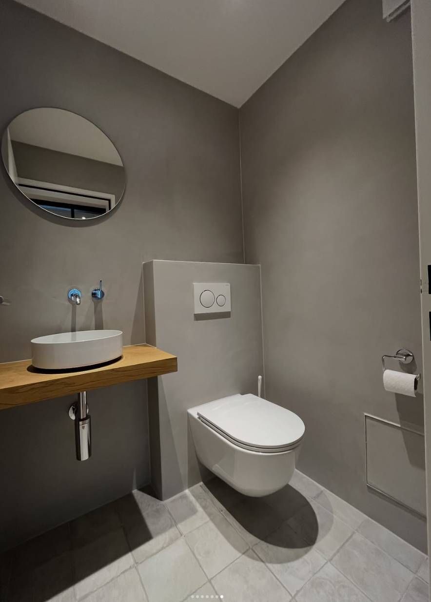 Ein modernes, minimalistisches Gäste-WC mit grauen Wänden, wandhängender Toilette, einem Waschtischregal aus Holz und einem runden Wandspiegel.