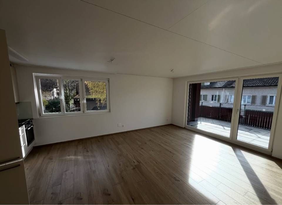 Ein leerer Raum mit hellem Holzboden, weissen Wänden und Blick auf einen Balkon durch grosse Glastüren.