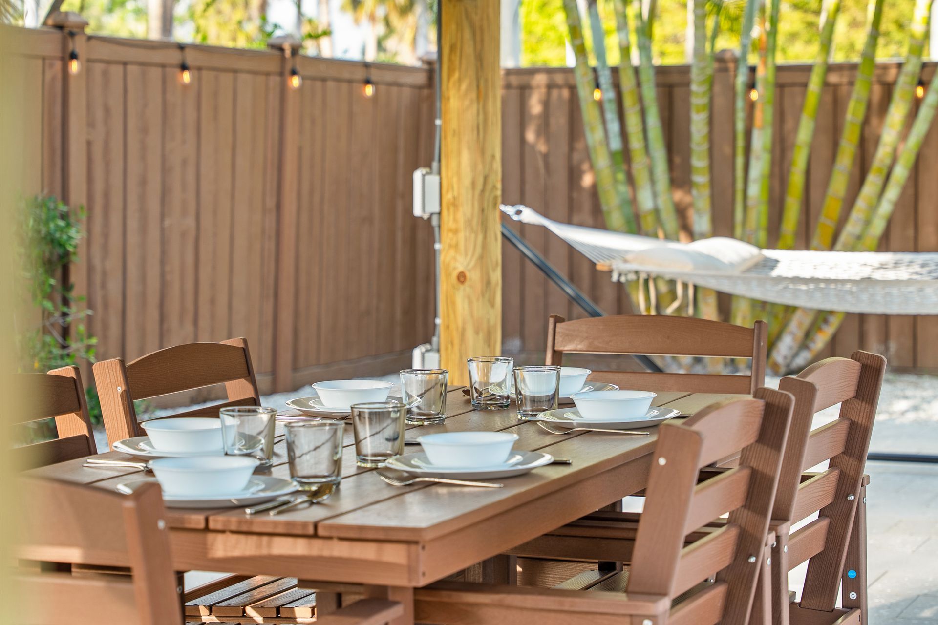 Outdoor patio dining table set with plates and glasses beside a hammock, fenced yard in the background