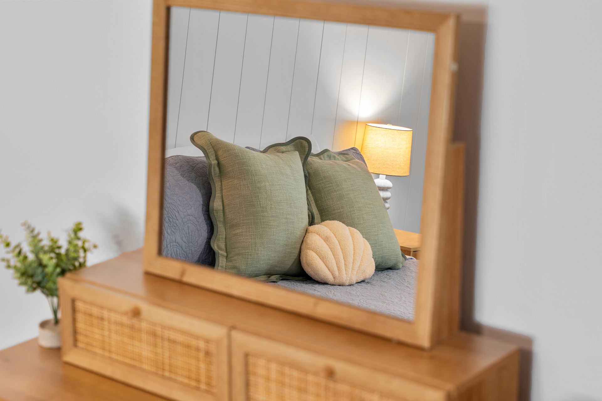 Wooden-framed mirror reflecting a bed with green pillows and a lit bedside lamp on a dresser