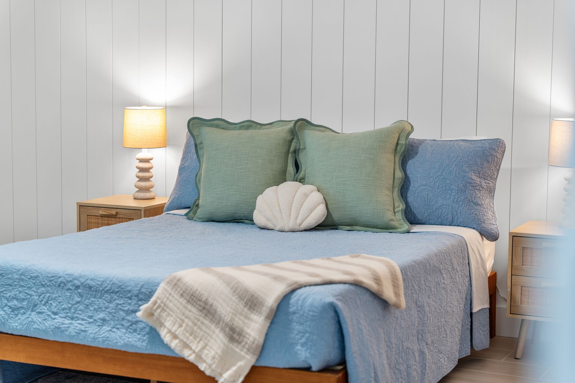 Blue bed with layered pillows and a folded blanket in a bright bedroom with bedside lamps