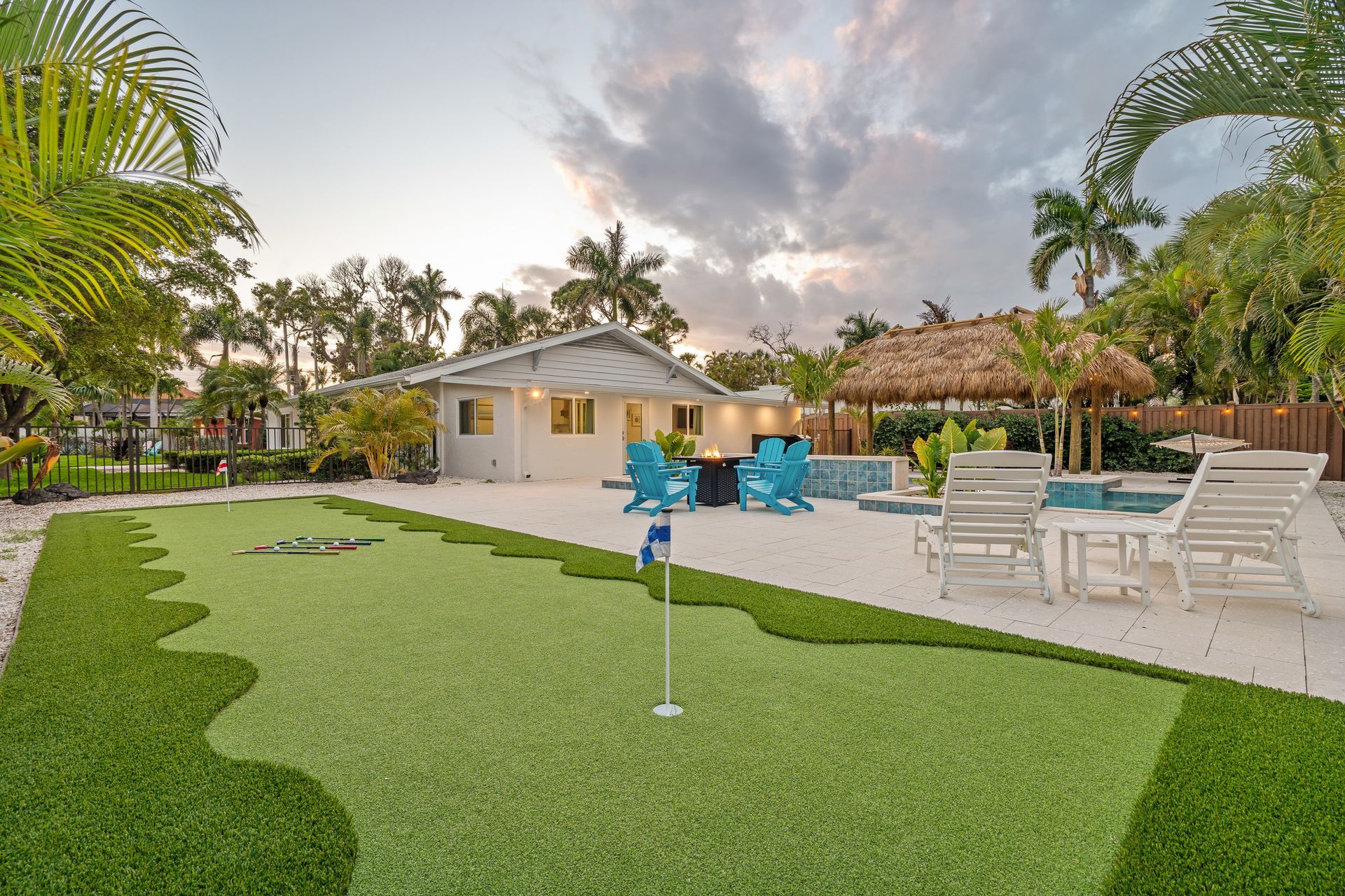 Backyard patio with pool, white lounge chairs, and a small putting green beside a tropical garden
