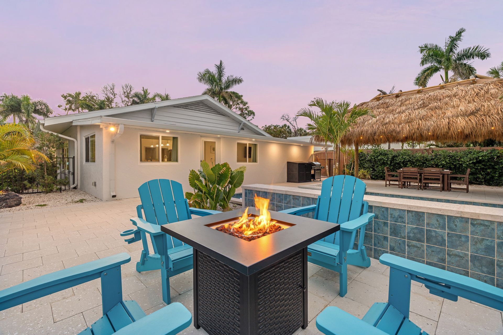 Tropical patio with blue chairs around a fire pit beside a pool at dusk