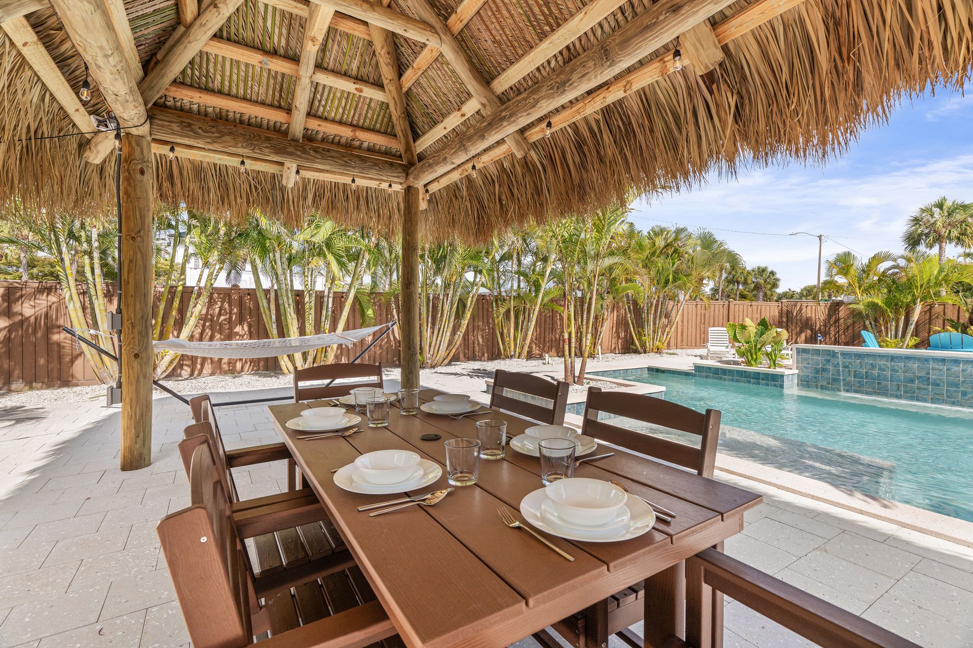 Thatched poolside dining area with wooden table, white dishes, and turquoise resort pool beside it