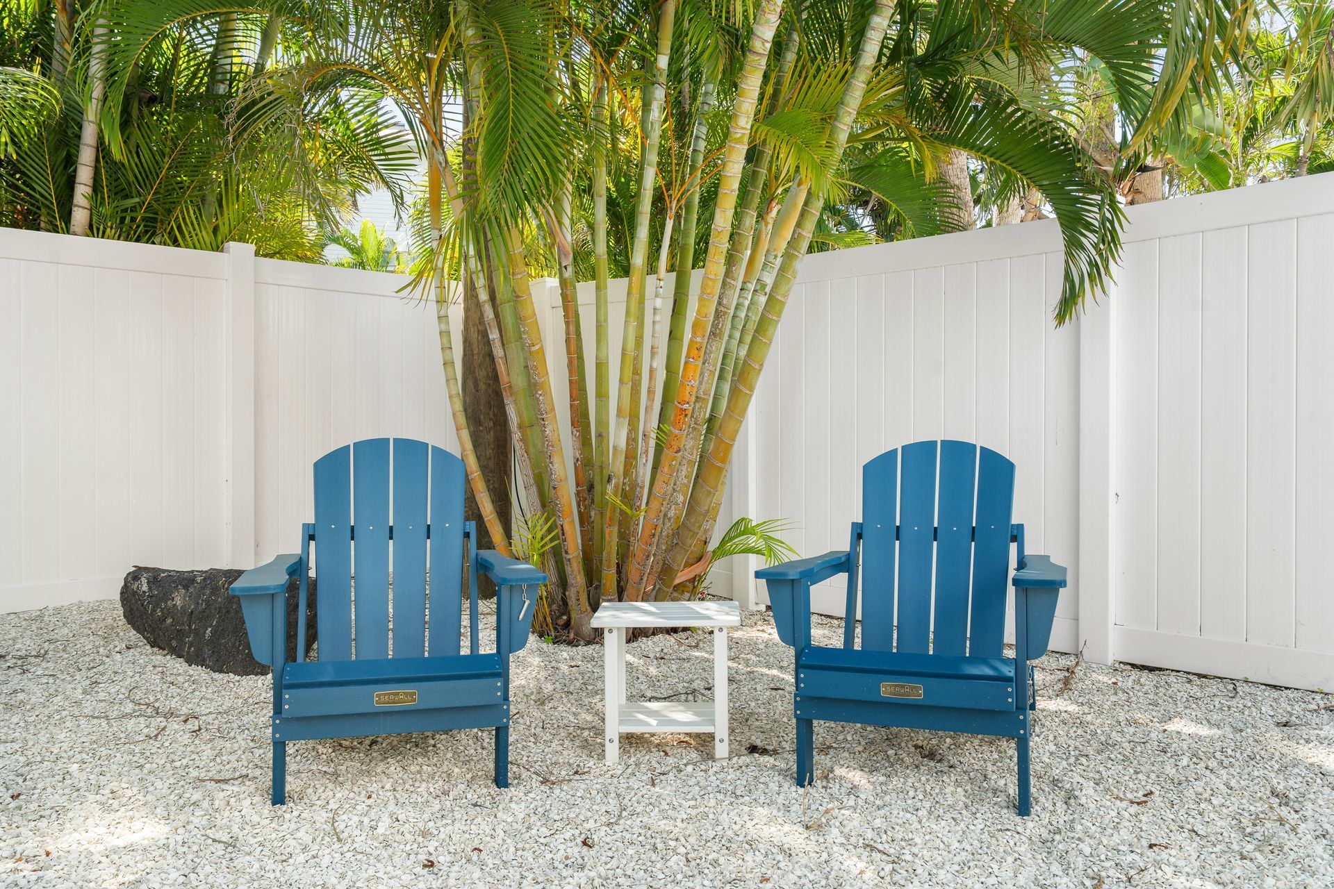 Two blue Adirondack chairs around a small white table in a tropical backyard courtyard