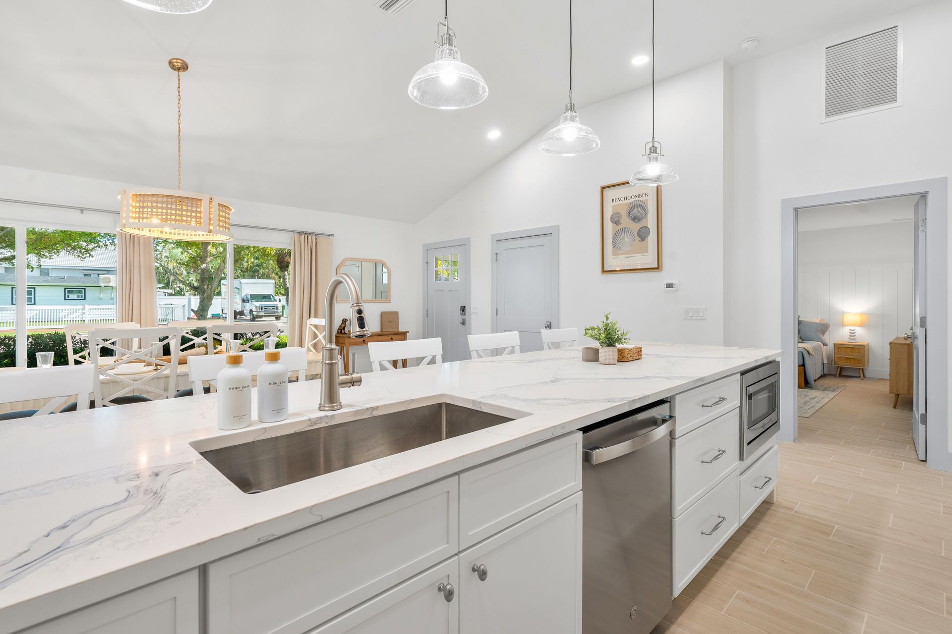 Bright white modern kitchen with island, stainless sink, pendant lights, and view into a dining area.