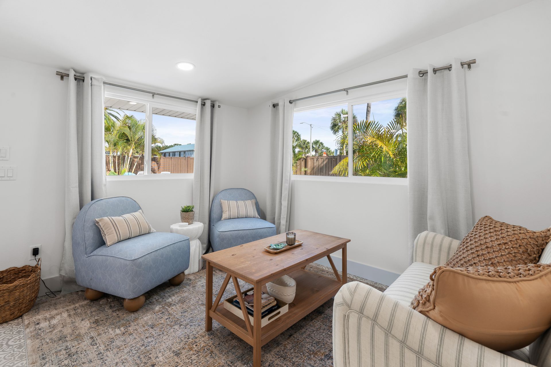 Bright living room with two blue chairs, white sofa, wooden coffee table, and large windows.