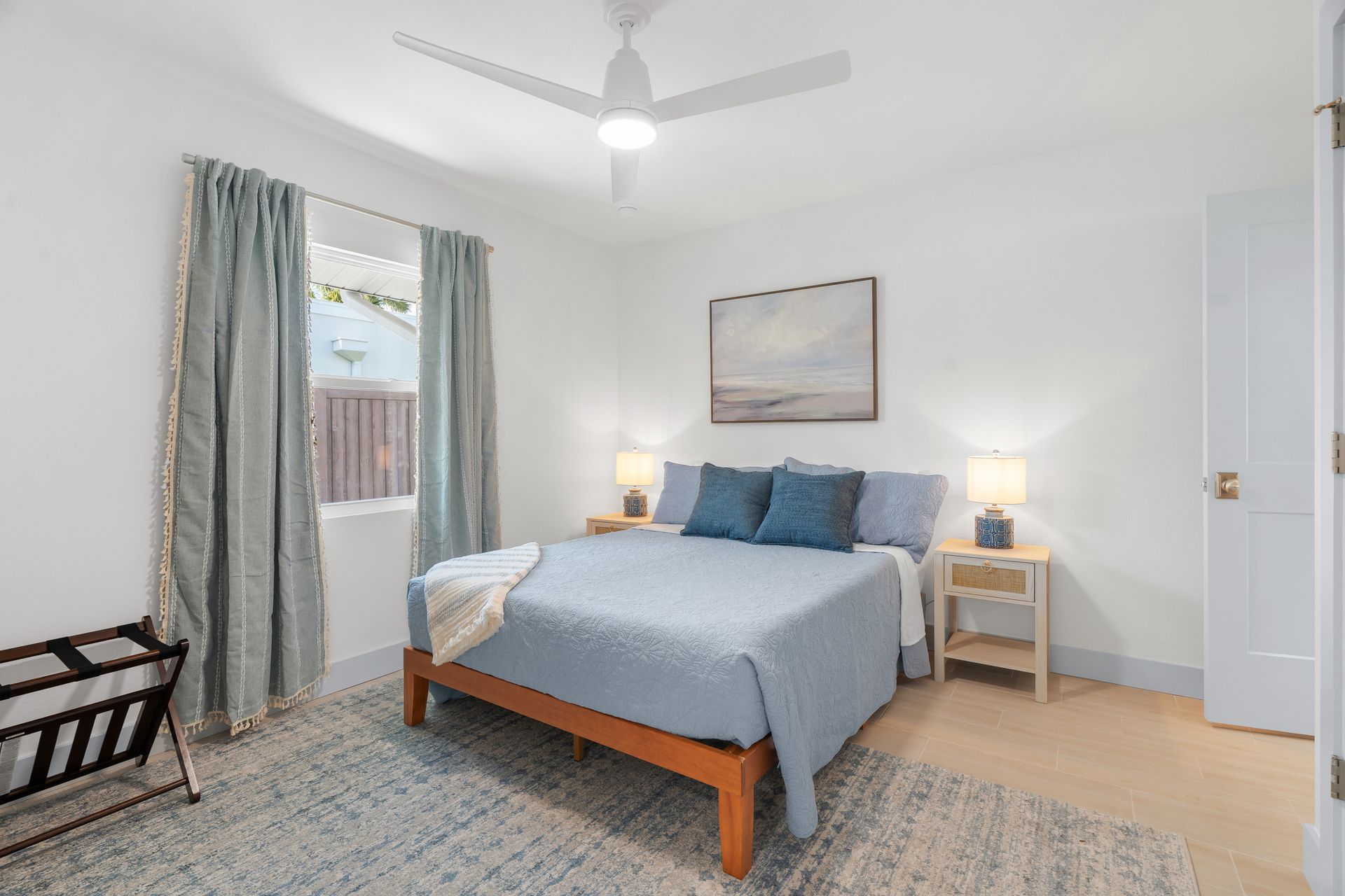 Bright bedroom with a wooden bed, blue bedding, nightstands, lamp, window, and patterned rug