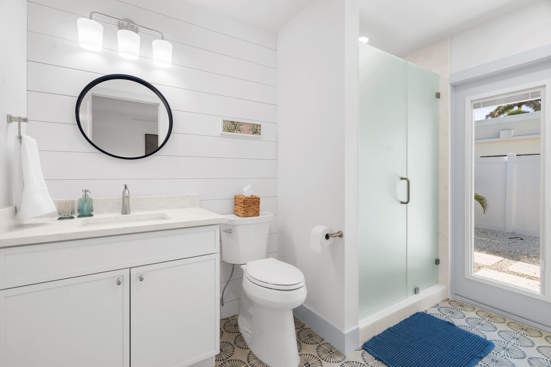 Bright modern bathroom with white vanity, round mirror, glass shower, and blue rug near a door