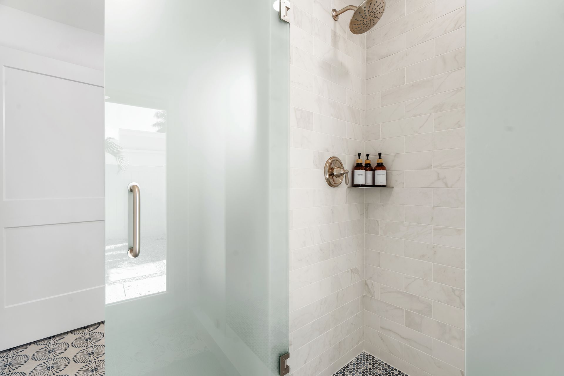 Modern white shower with glass door, built-in shelf of bottles, and pebble tile floor