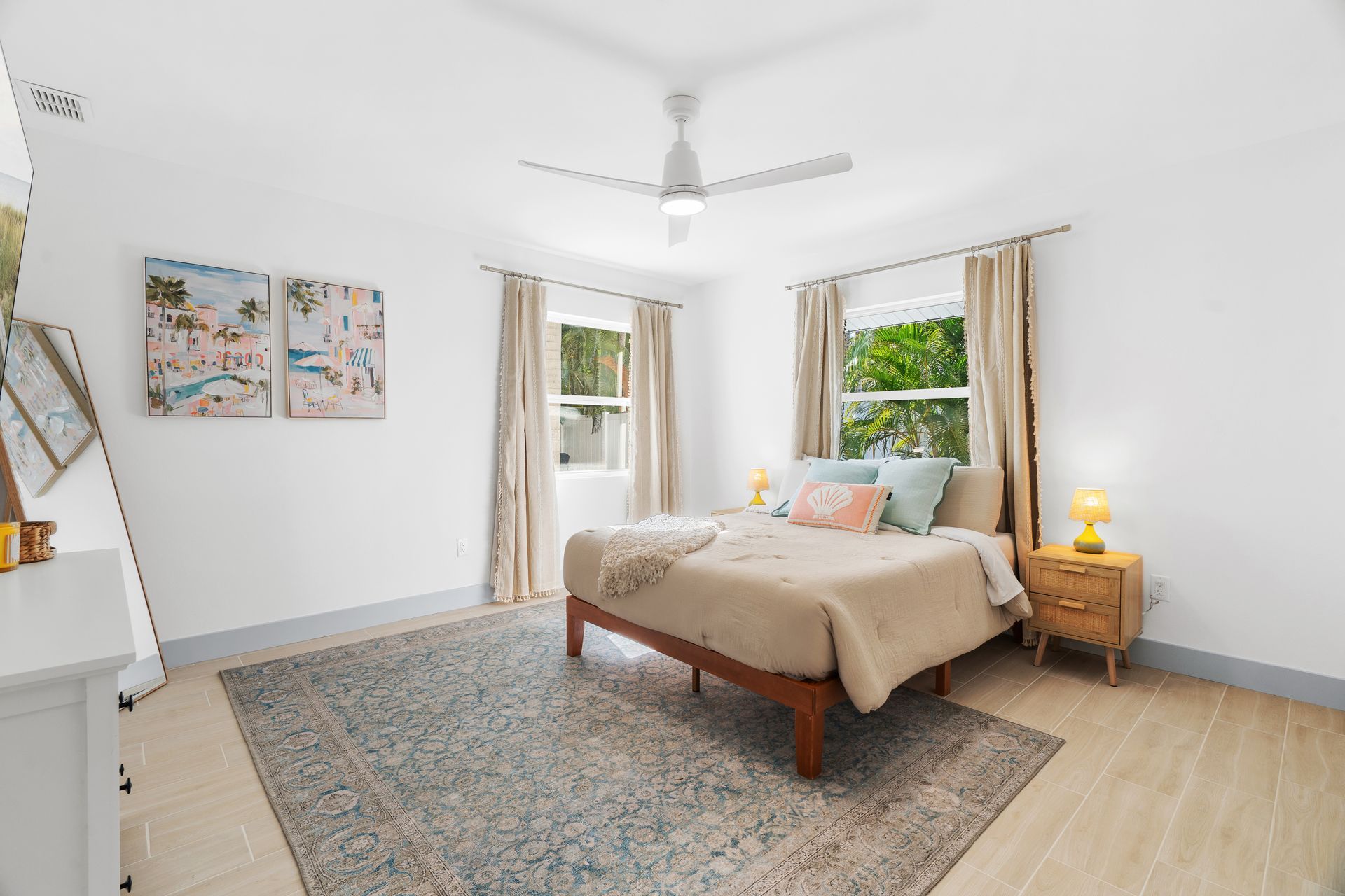 Bright bedroom with beige bed, patterned rug, two windows, ceiling fan, and white walls