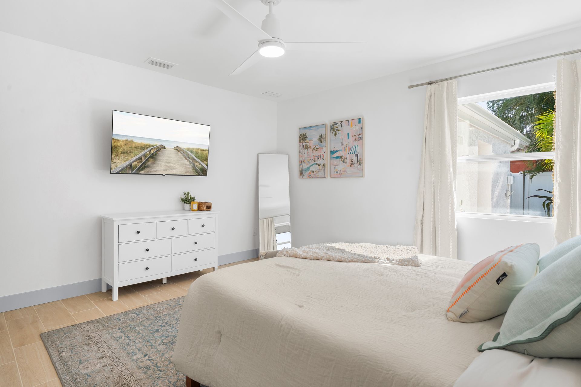 Bright bedroom with a bed, dresser, wall art, mirror, and large window with white curtains.