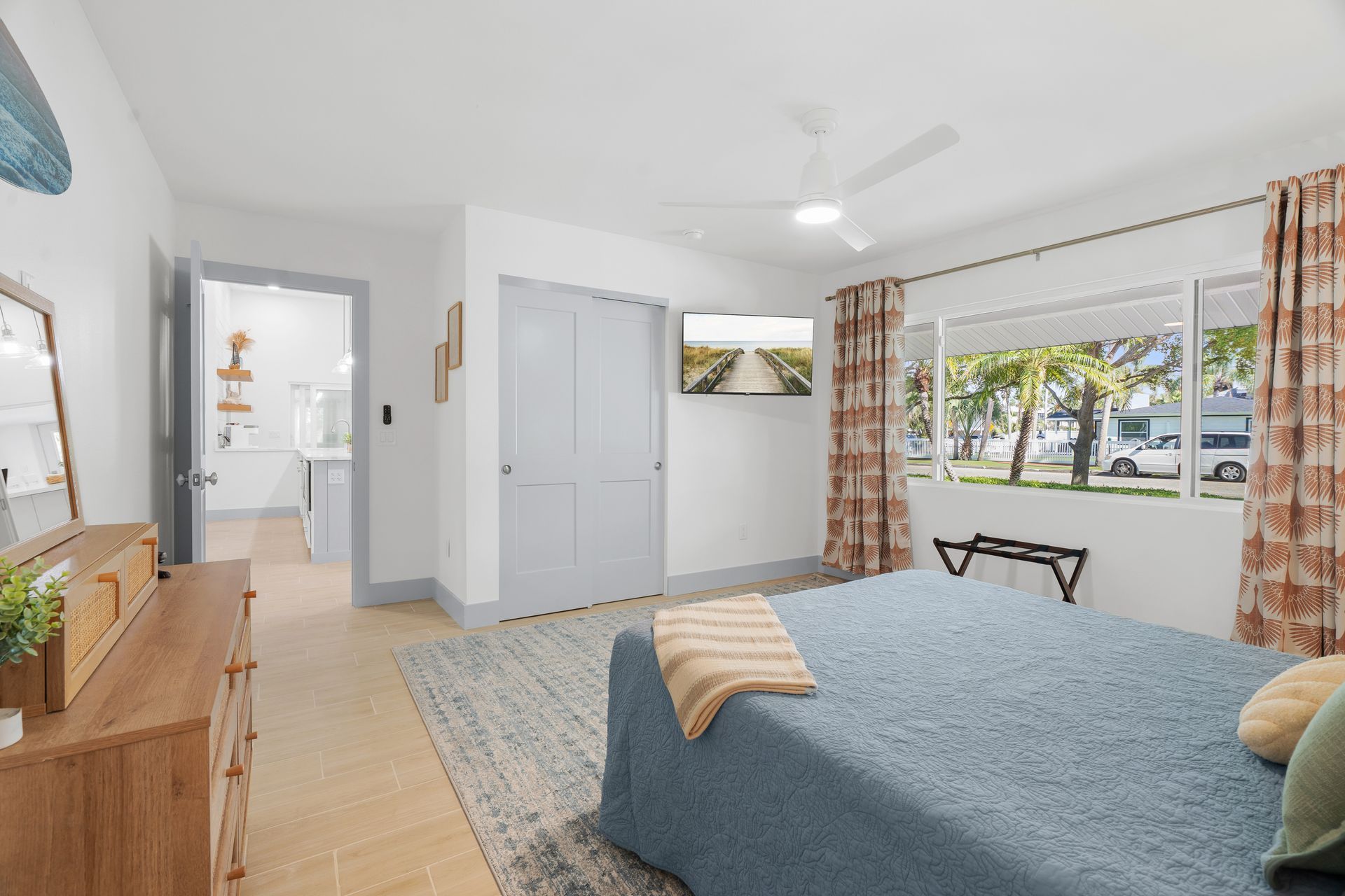 Bright bedroom with blue bedspread, white walls, dresser, closet, and large window overlooking trees and parked cars