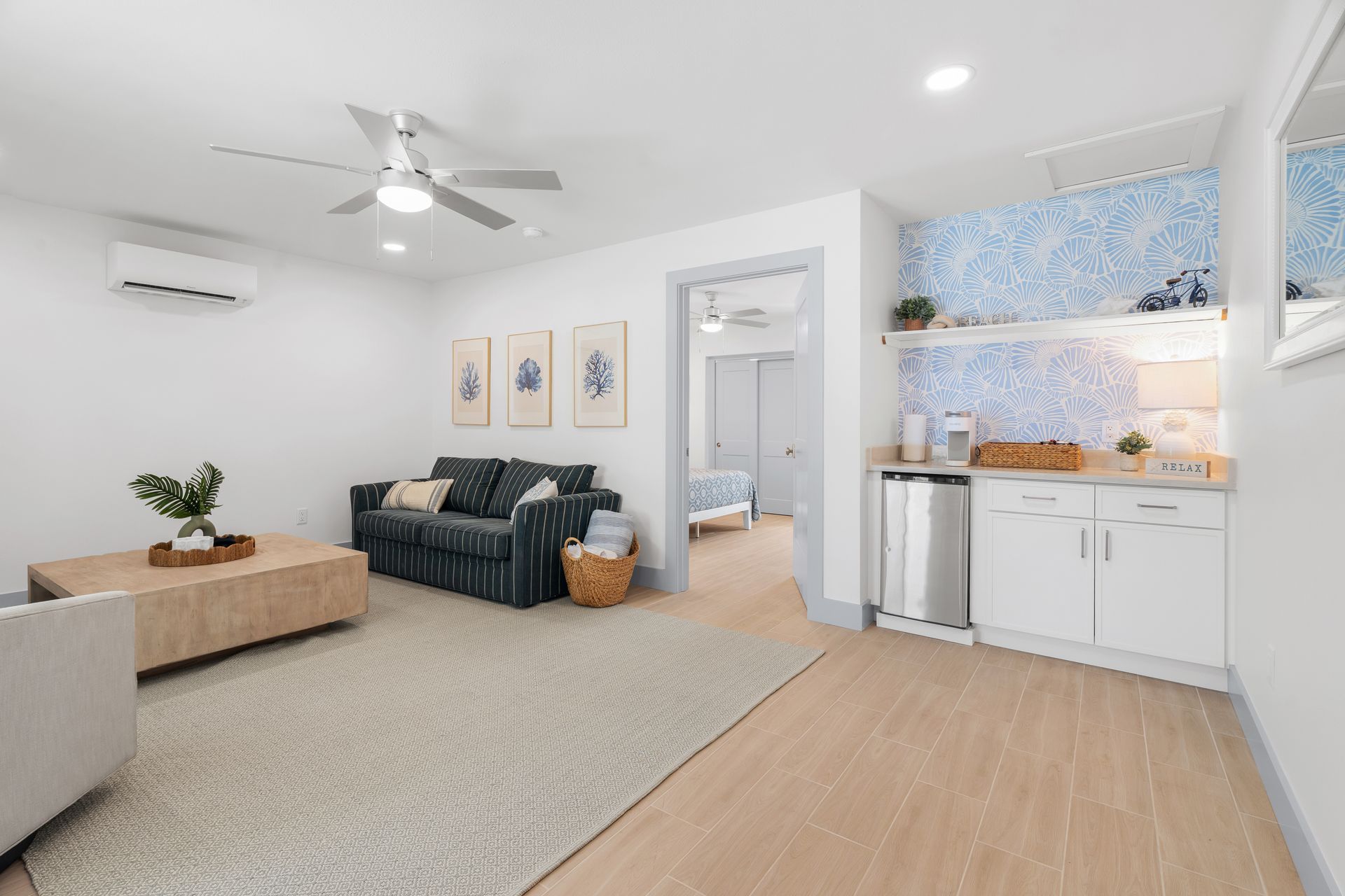 Bright open living room with gray sofa, wood coffee table, patterned rug, and a small kitchen area.