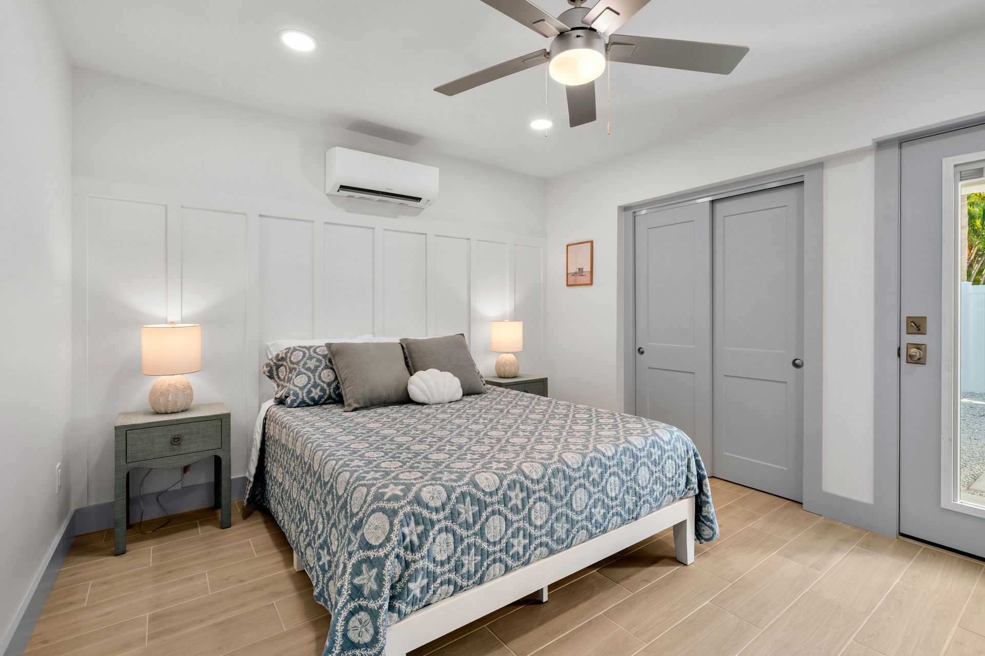 Bright bedroom with blue patterned bedspread, gray closet doors, nightstands, ceiling fan, and wood floor
