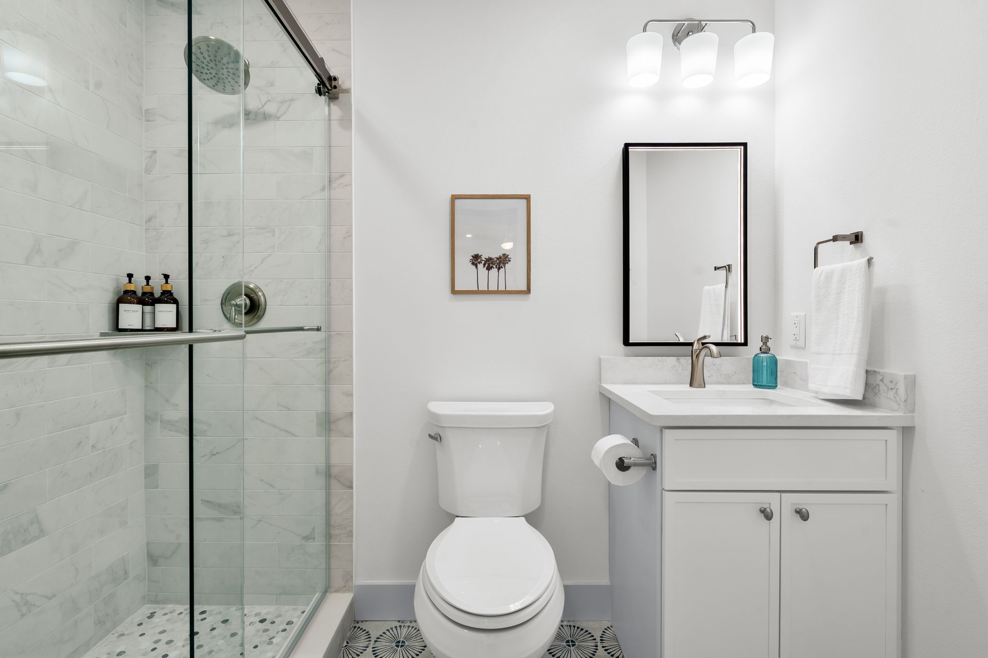 Bright white bathroom with glass shower, toilet, sink, and mirror on a light tile floor