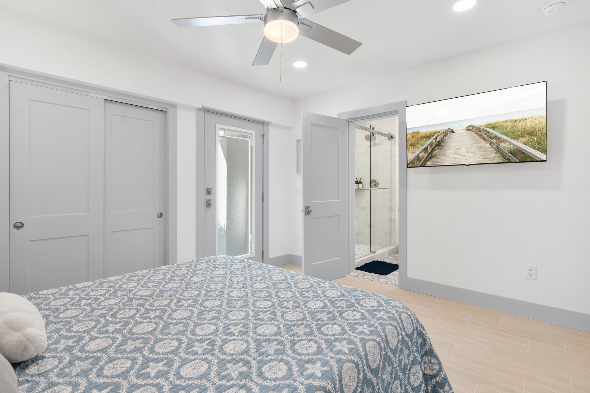 Bright bedroom with blue patterned bedspread, open doors, ceiling fan, and a wall-mounted landscape print.
