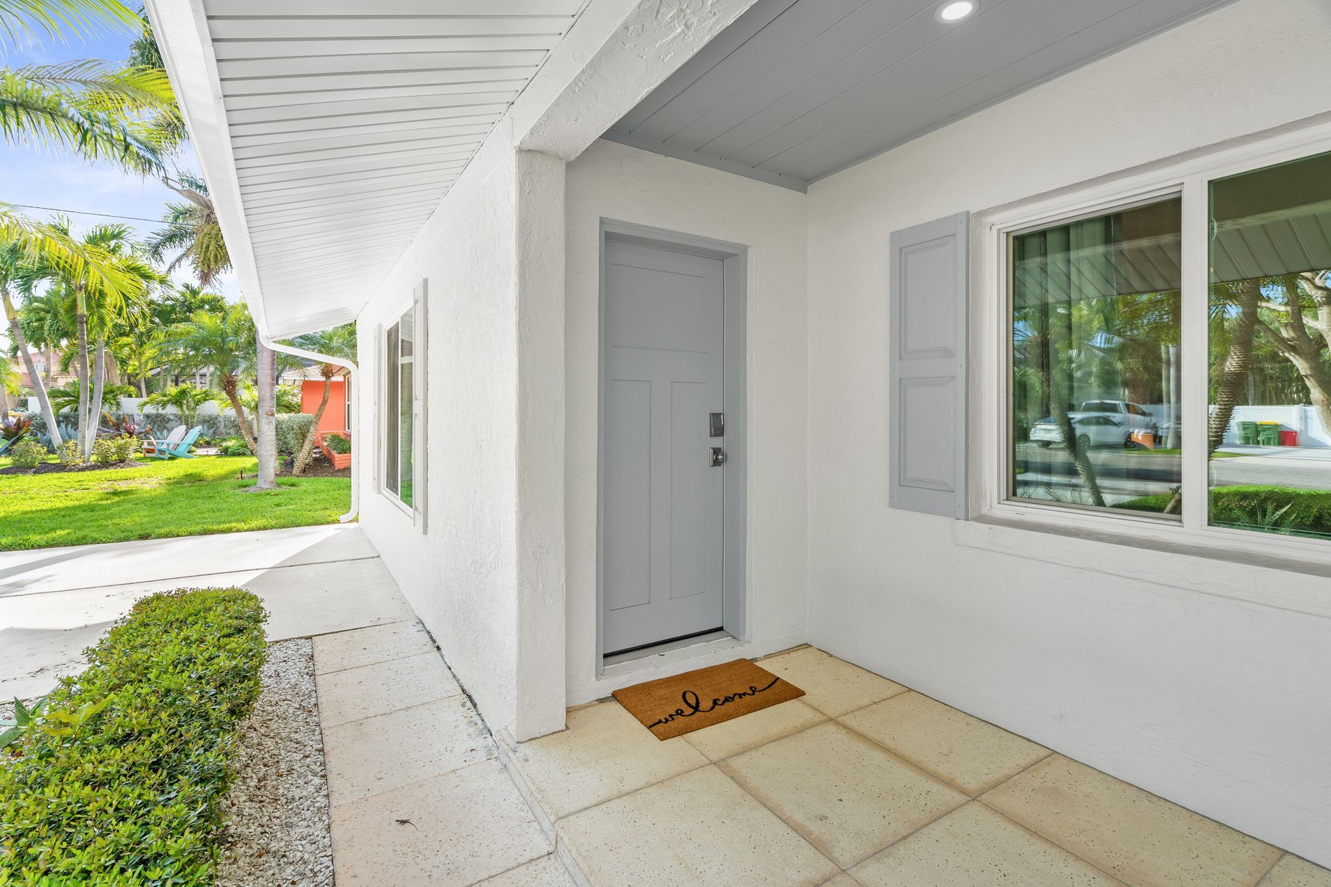 Covered front porch with white door, window, potted greenery, and a welcome mat beside a sidewalk.