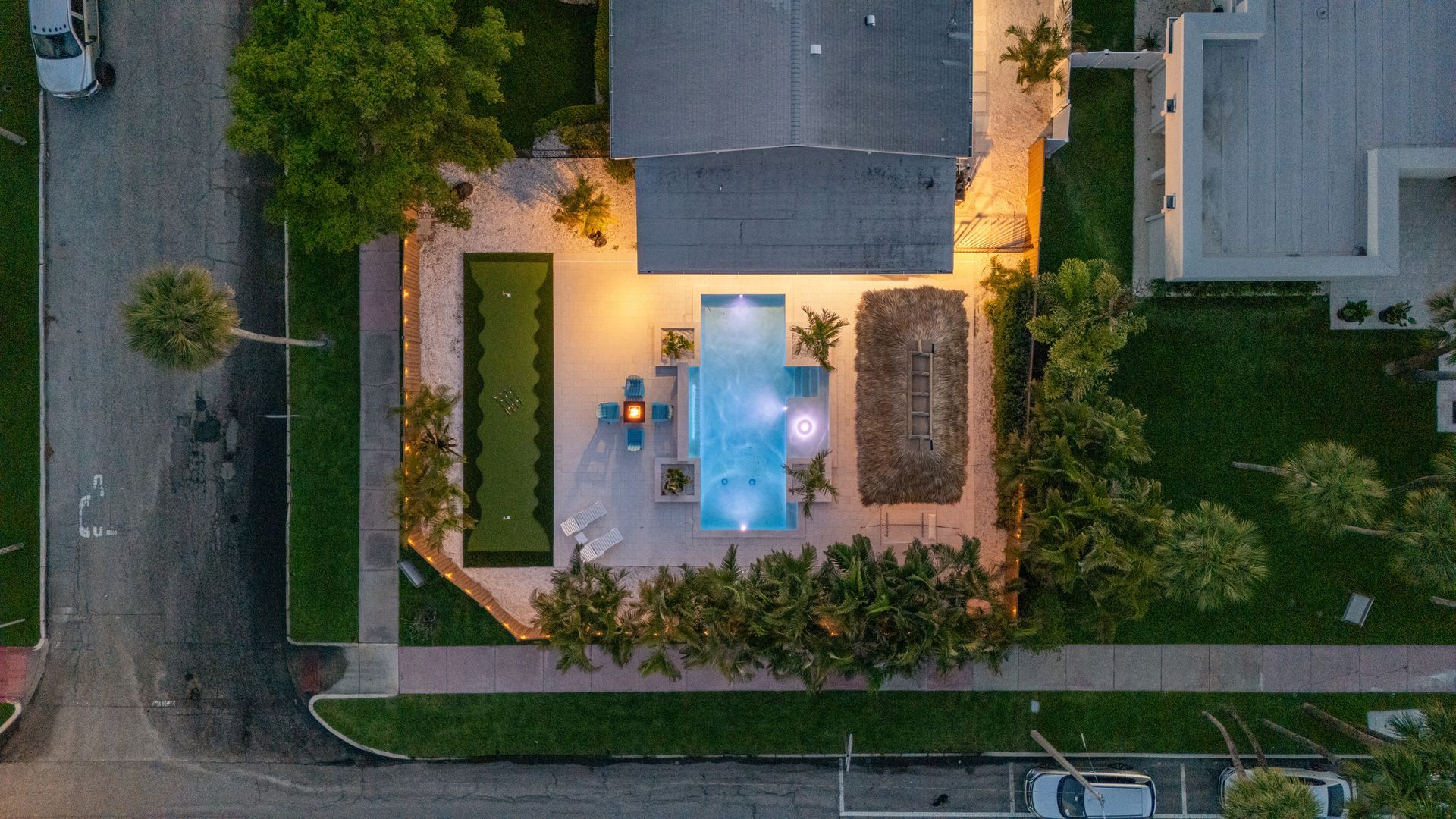 Aerial view of a lit backyard patio with a swimming pool, lounge chairs, and surrounding greenery at dusk