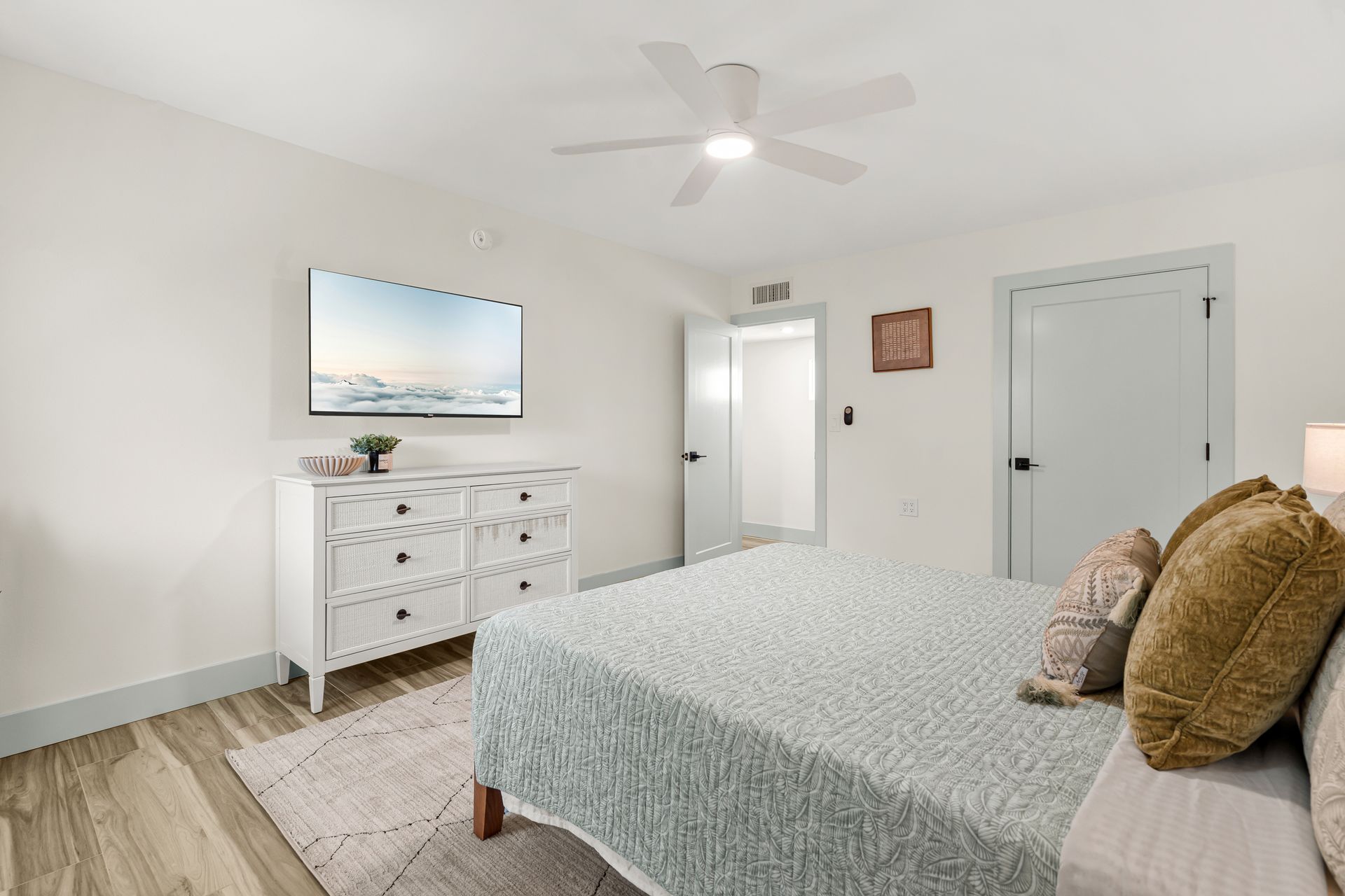 Bright bedroom with white dresser, bed, ceiling fan, and two closed doors on light wood floors.