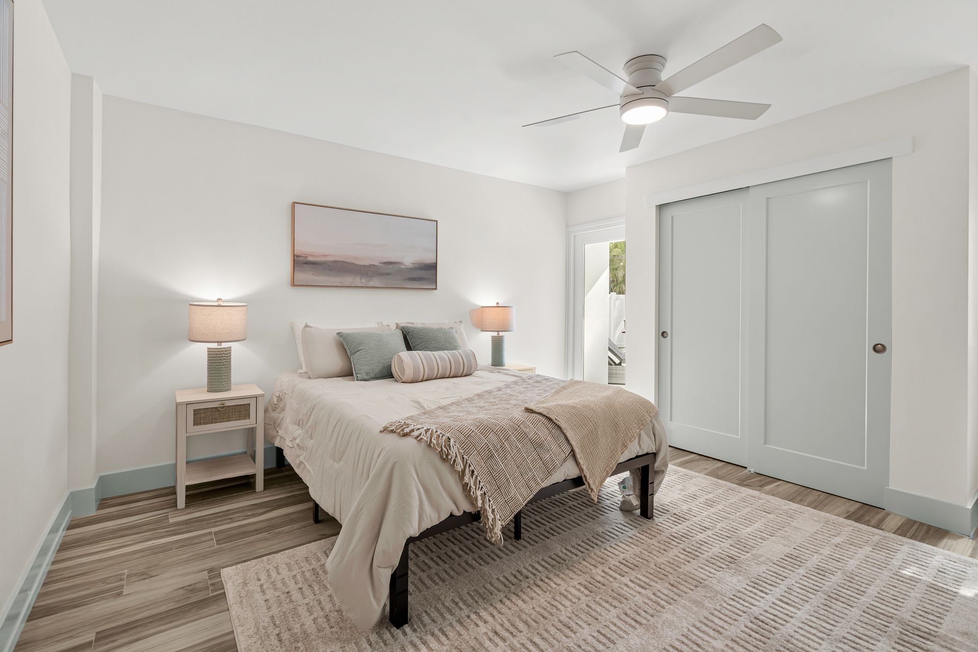 Bright bedroom with a bed, nightstands, lamps, ceiling fan, and closet doors on a light rug.