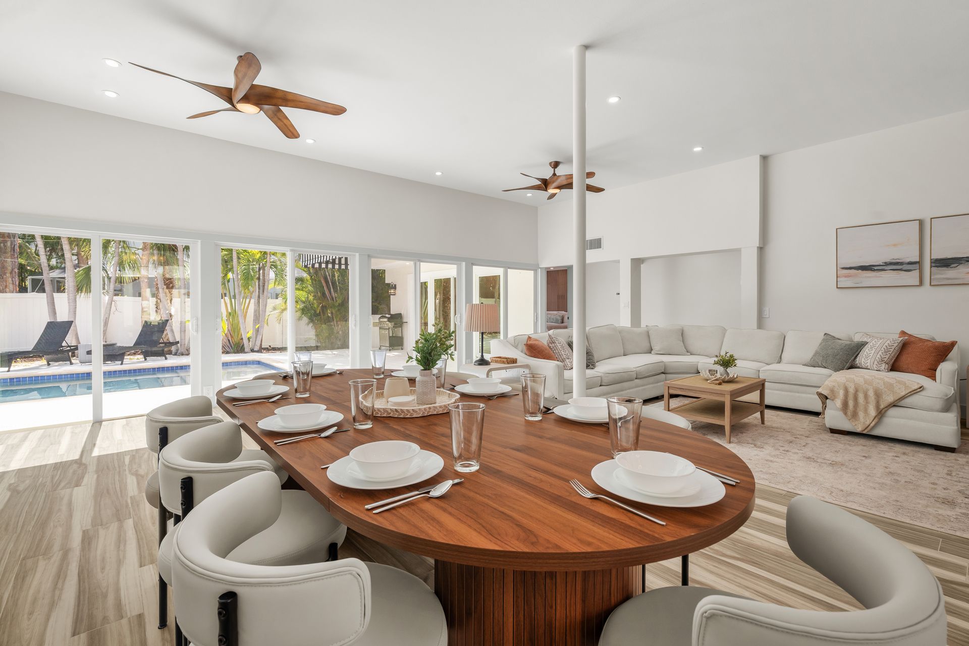 Bright open dining room with oval wooden table, white chairs, ceiling fans, and sliding doors to a pool.